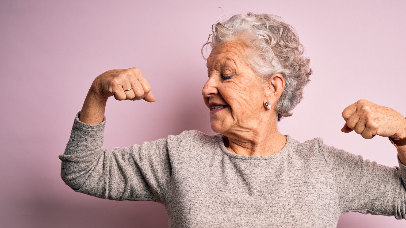 Senior beautiful woman wearing casual t-shirt standing over isolated pink background showing arms muscles smiling proud. Fitness concept.